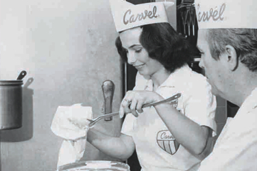 This is a black and white photograph featuring two individuals in a kitchen setting. The person on the left appears to be a woman wearing a uniform that includes a hat, apron, and badge; she has her hands placed on the countertop, with one hand holding an object wrapped in white paper. In the center of the image is a man dressed in a chef's outfit, standing behind the woman with his attention focused towards her. Both individuals are engaging in culinary activities. The woman is near what appears to be a mixing bowl, while the man is near what might be a frying pan or griddle. There is a cake in front of them, which has been partially decorated and sits on a flat surface. In the background, there are various kitchen utensils visible, including a roll of white paper, multiple spoons, and a baking dish containing a golden-brown substance. The setting suggests that this could be a snapshot taken during a cooking activity or showcase event.